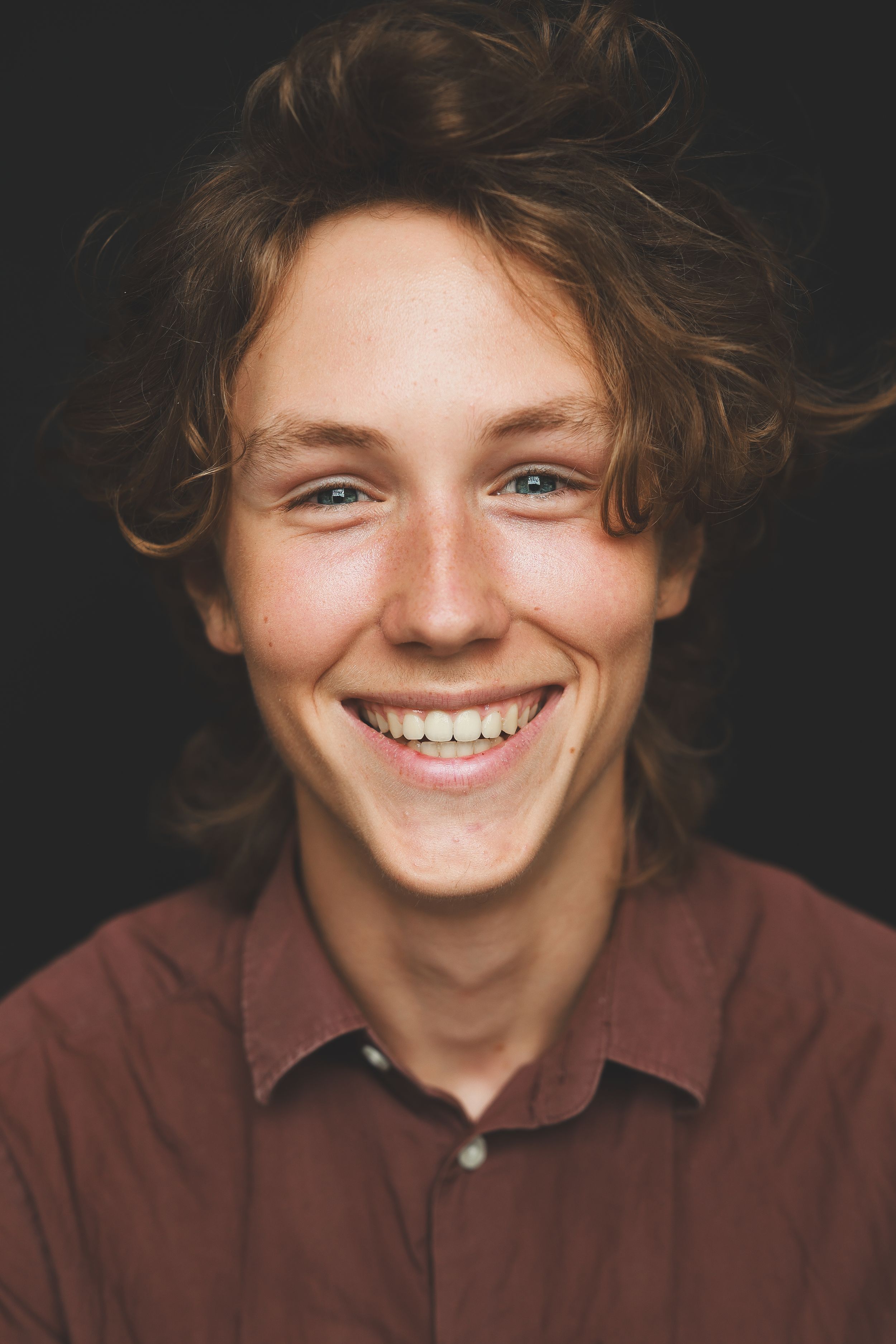 Happy teenage boy with curly auburn hair and blue eyes smiling in a burgundy shirt against dark background.