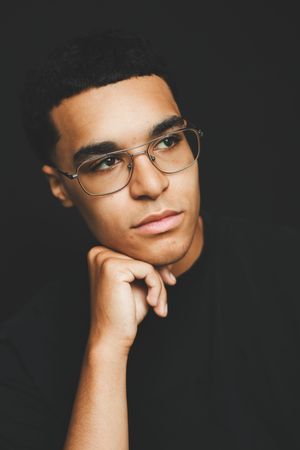 Thoughtful young man with glasses resting chin on hand wearing a black top against dark background.