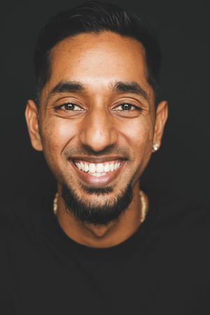 Smiling young man with beard and stud earring wearing a black top against a dark background.