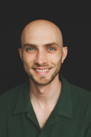 Bald young man with blue eyes and short beard smiling in a green button-up shirt against dark background.