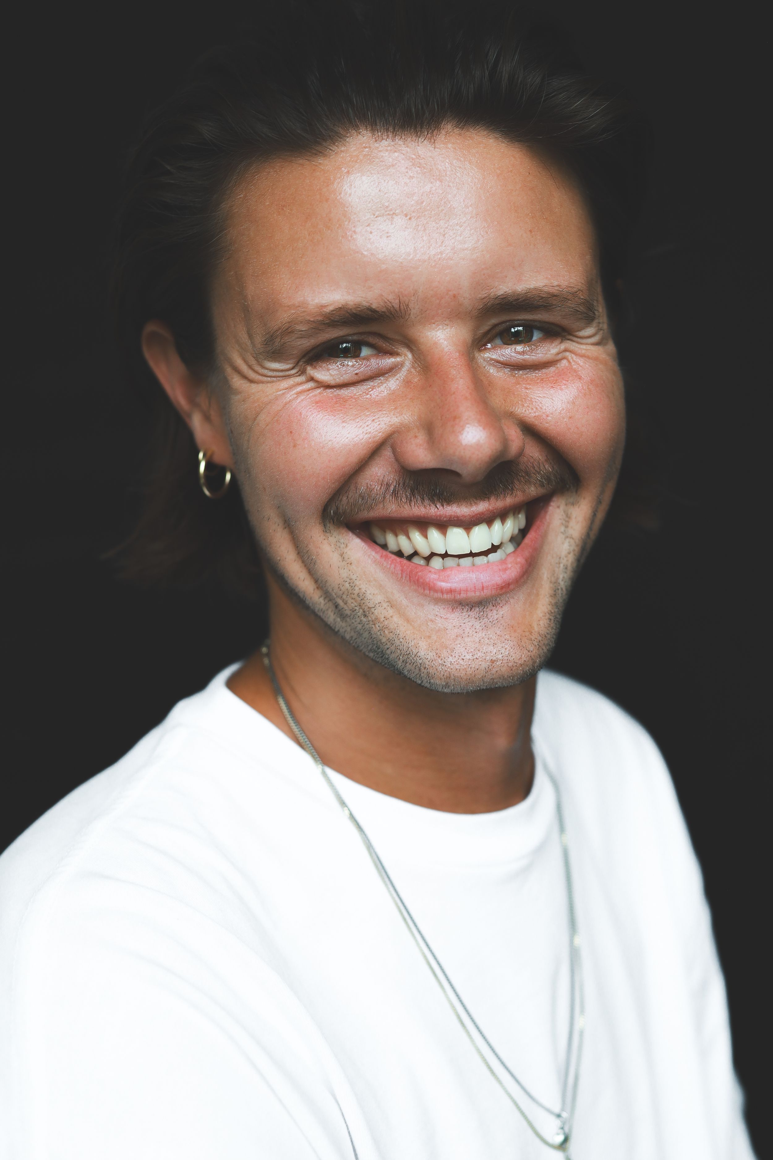 Happy young man with dark swept-back hair and hoop earring laughing in a white t-shirt against dark background.