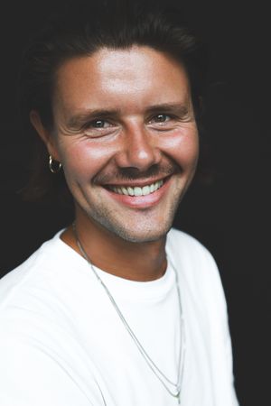 Smiling young man with dark hair, hoop earring, and silver necklace wearing a white t-shirt against dark background.