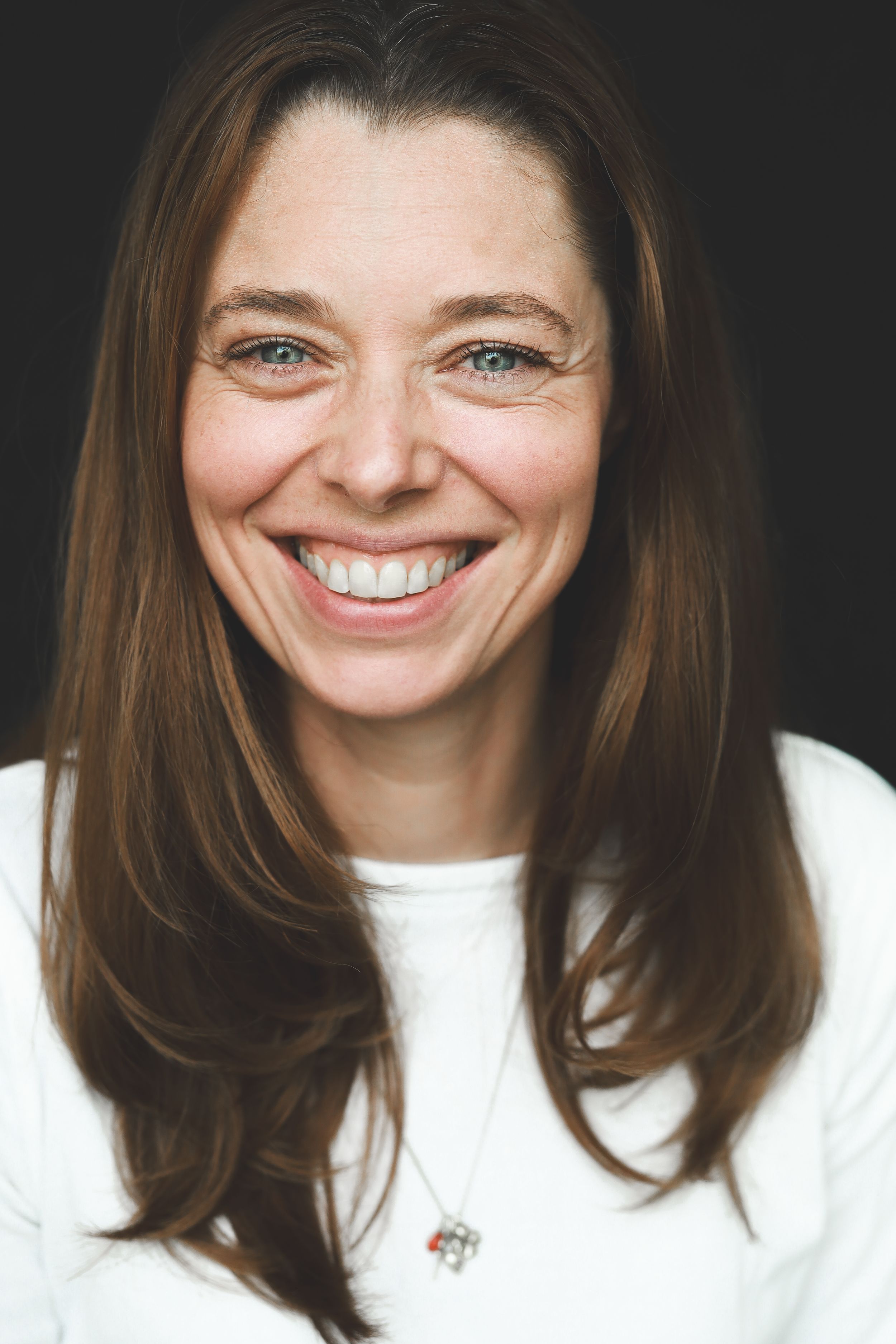 Portrait of a smiling brunette woman with blue eyes and long hair wearing a white top and necklace.