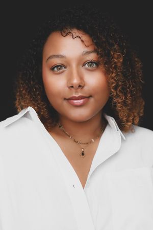 Portrait of a young woman with curly auburn hair and green eyes wearing a white shirt and gold necklace.