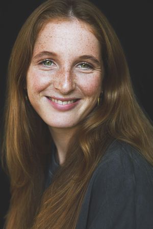Portrait of a smiling young woman with long red hair, freckles, and green eyes on a dark background.