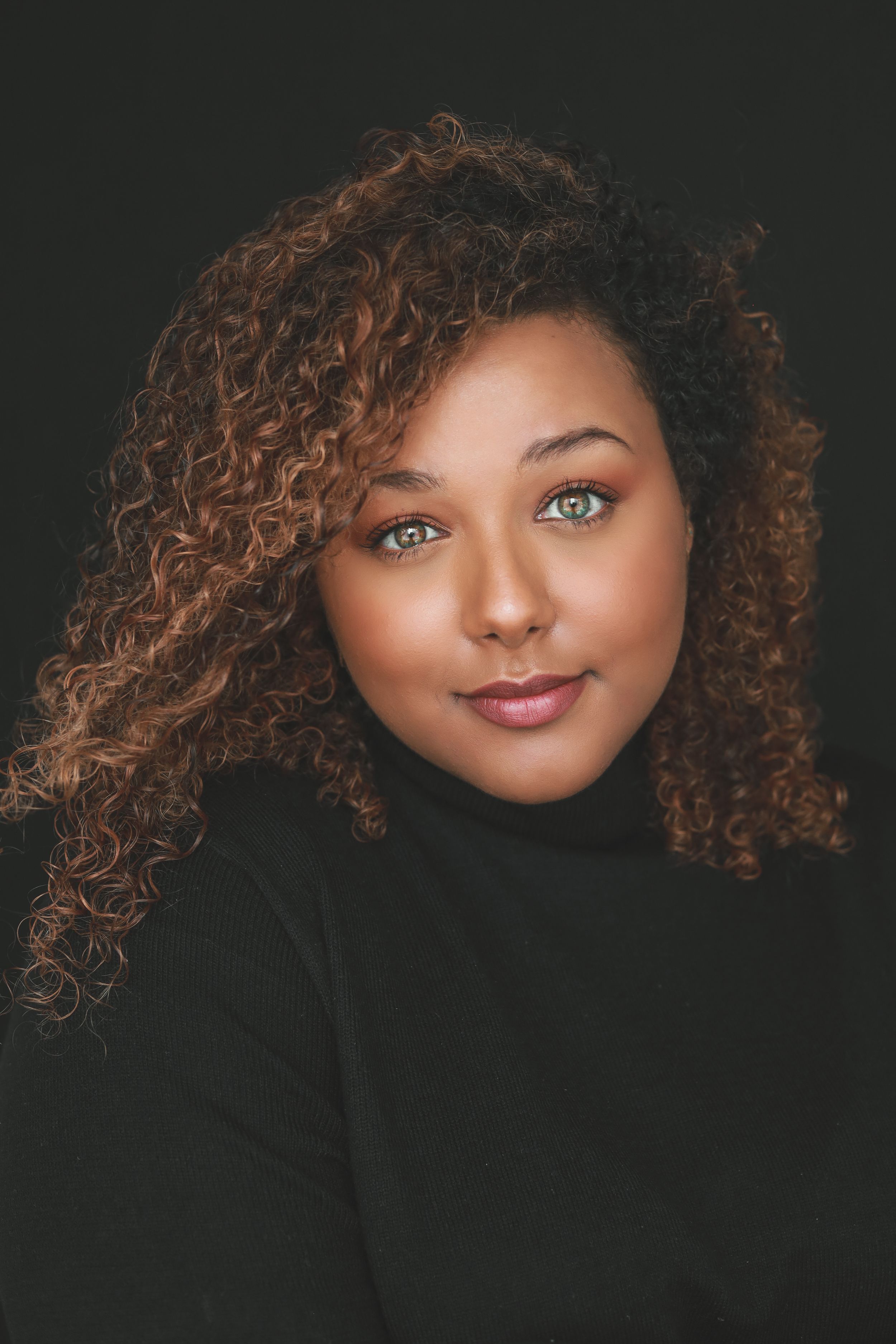 Young woman with curly auburn hair and green eyes wearing a black turtleneck against a dark background.