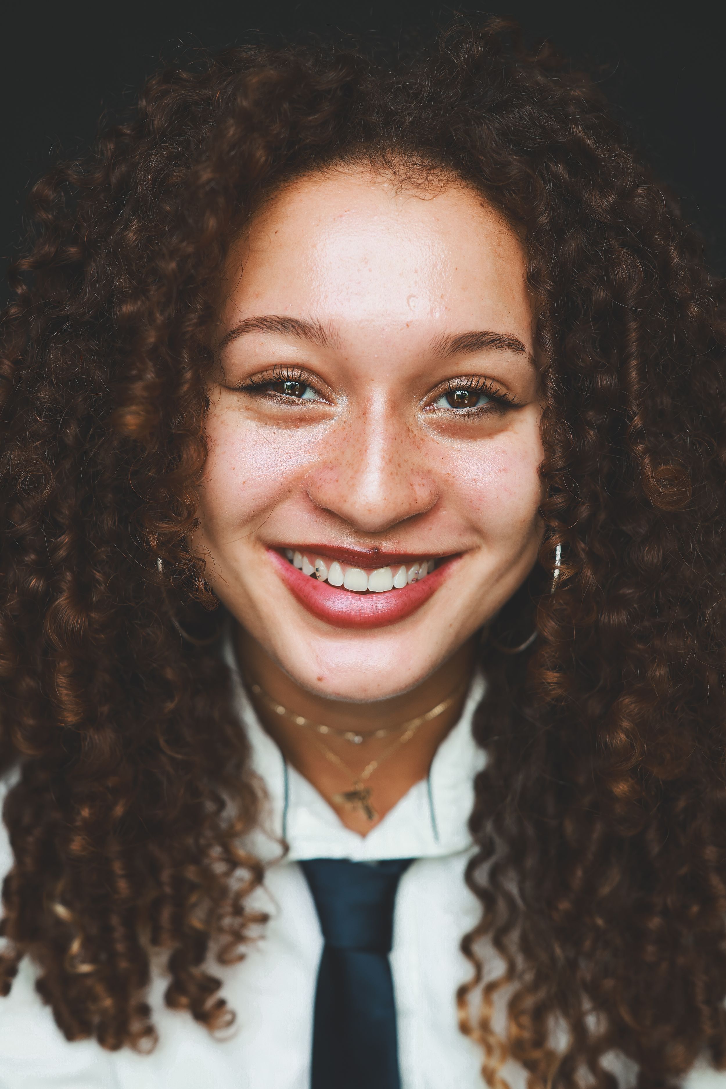 Cheerful young woman with curly hair, freckles, and hoop earrings wearing a white shirt and tie.