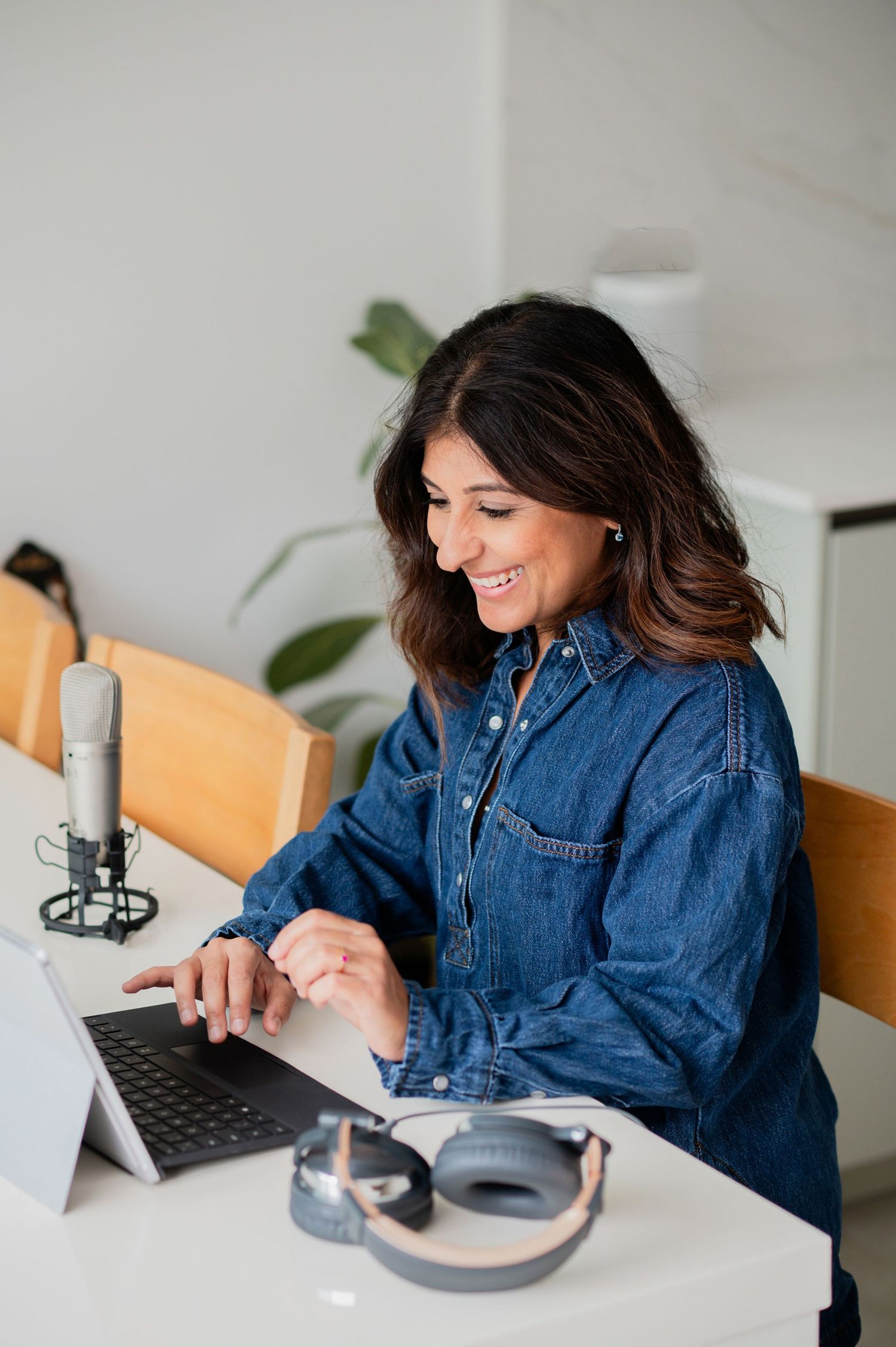 Smiling woman in denim shirt working on laptop with microphone and headphones on white desk.