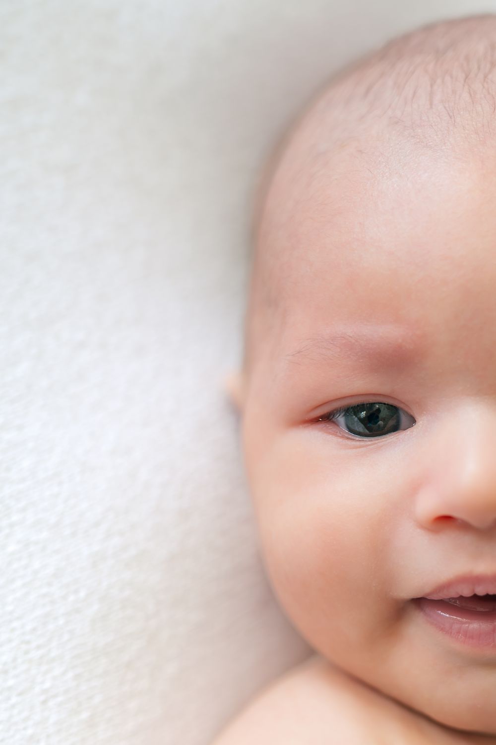 Close-up portrait of a smiling newborn baby with blue-gray eyes lying on a soft white blanket.