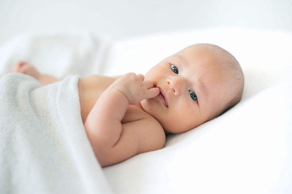 Adorable newborn baby lying on white blanket, fingers in mouth, with bright curious eyes and soft skin.