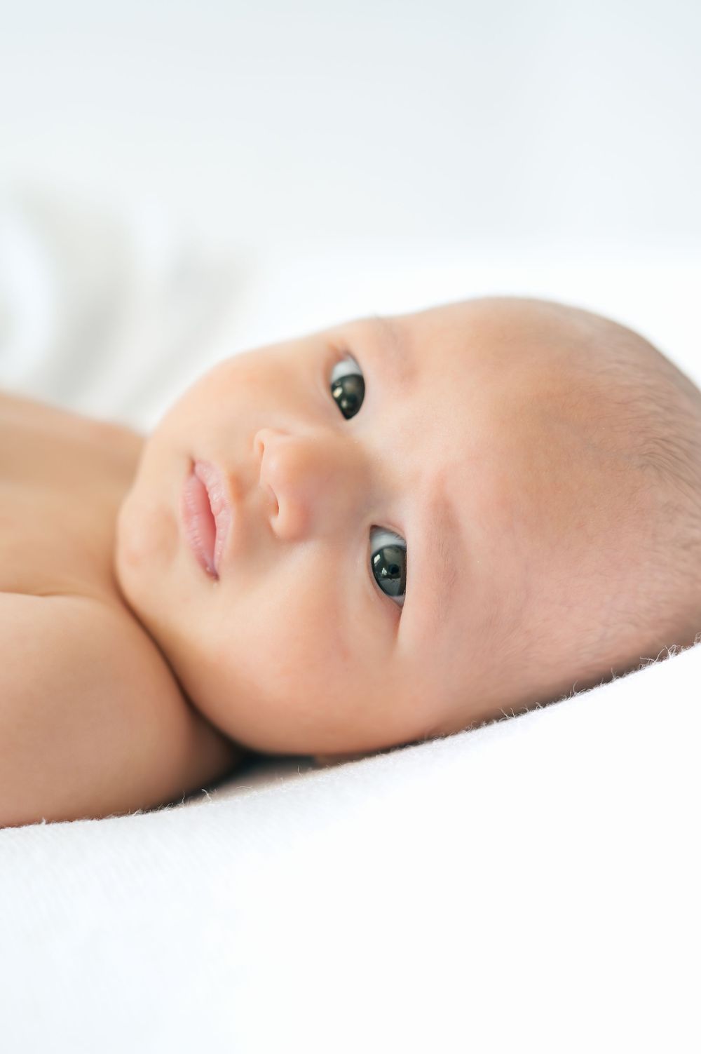 Close-up portrait of a newborn baby with dark eyes lying on a soft white blanket.