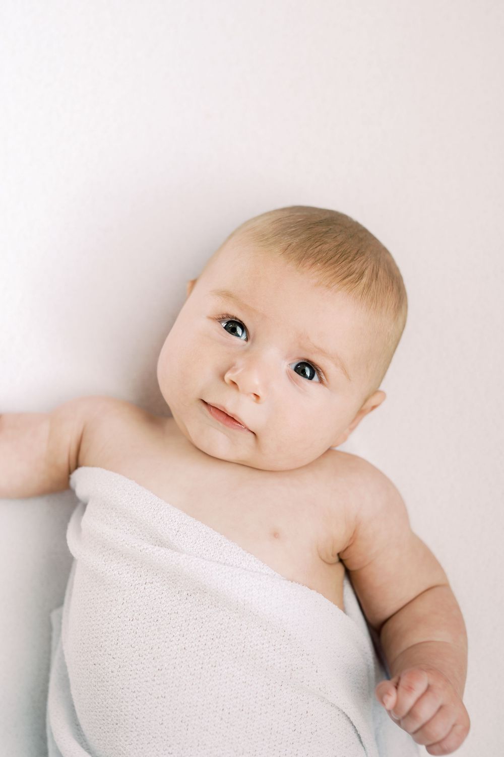 Adorable newborn baby wrapped in a soft white blanket, with blue eyes and light hair, against a white background.