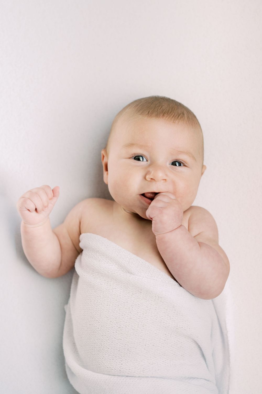 Adorable baby wrapped in white blanket lying down, chewing fingers with one fist raised, looking at camera.