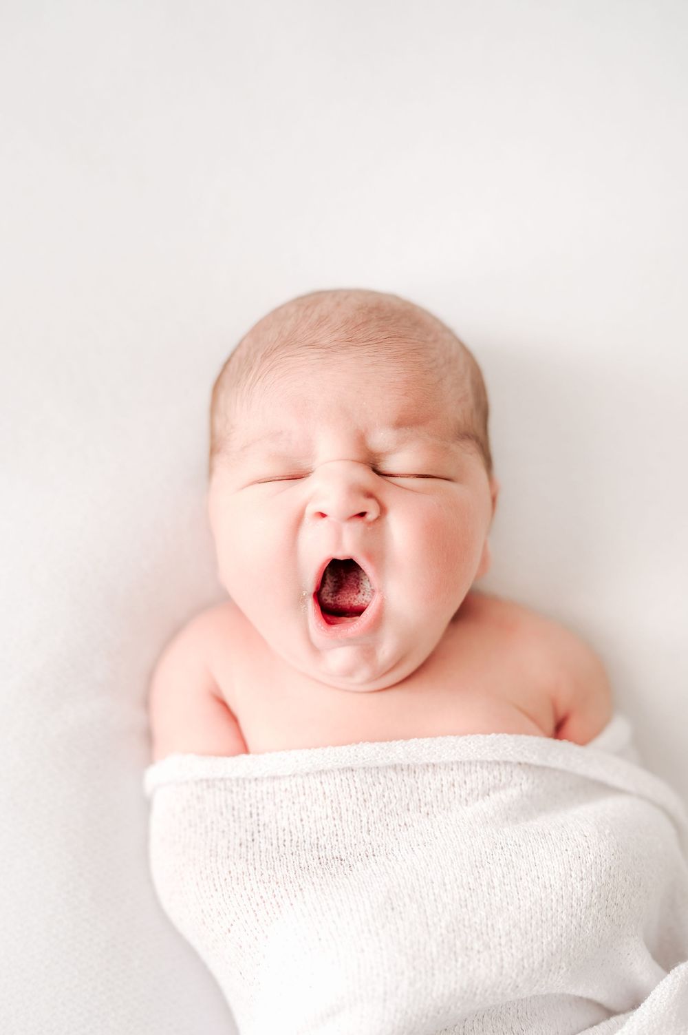 Newborn baby yawning while swaddled in a white blanket, lying on a white background.