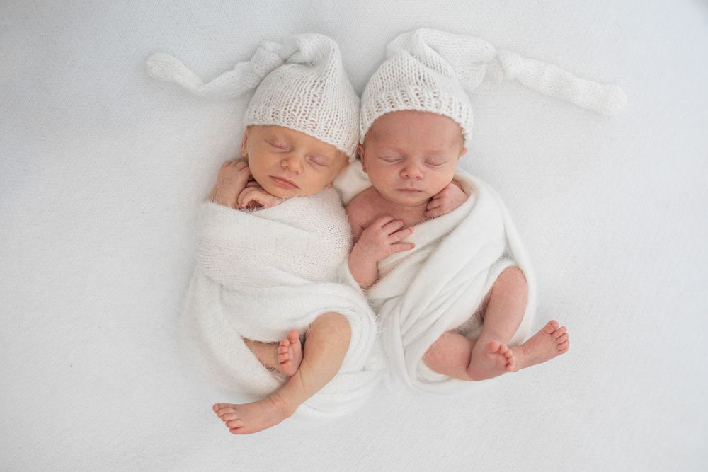 Two sleeping newborn twins wrapped in white swaddles wearing knit hats on a white background.