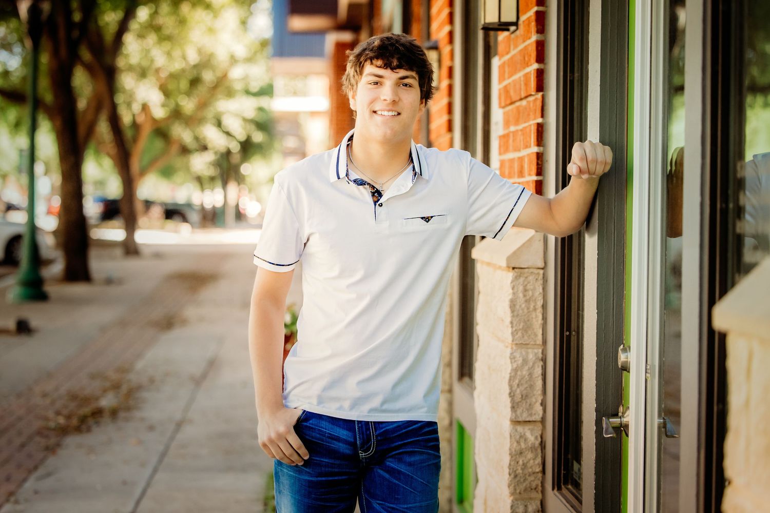 Young man in white polo and blue jeans smiling on a sunny small-town sidewalk near a storefront in Keller, TX.