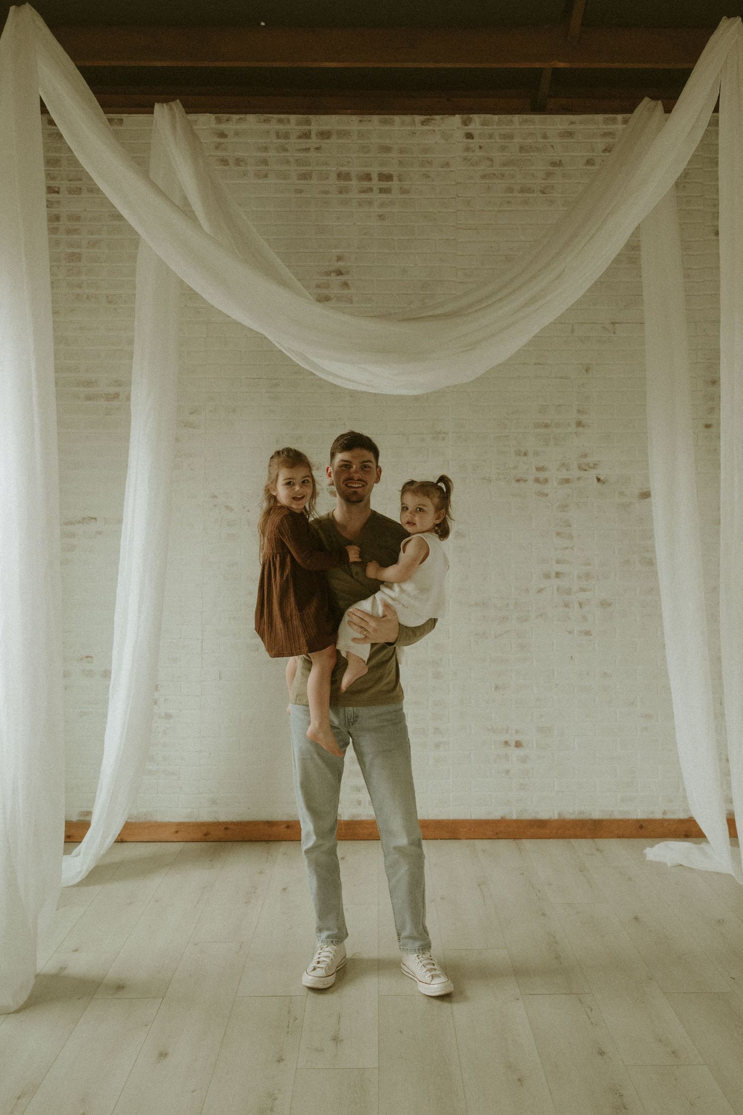 A parent and two children pose in front of a white brick wall with flowing fabric draping overhead in a minimalist setting.
