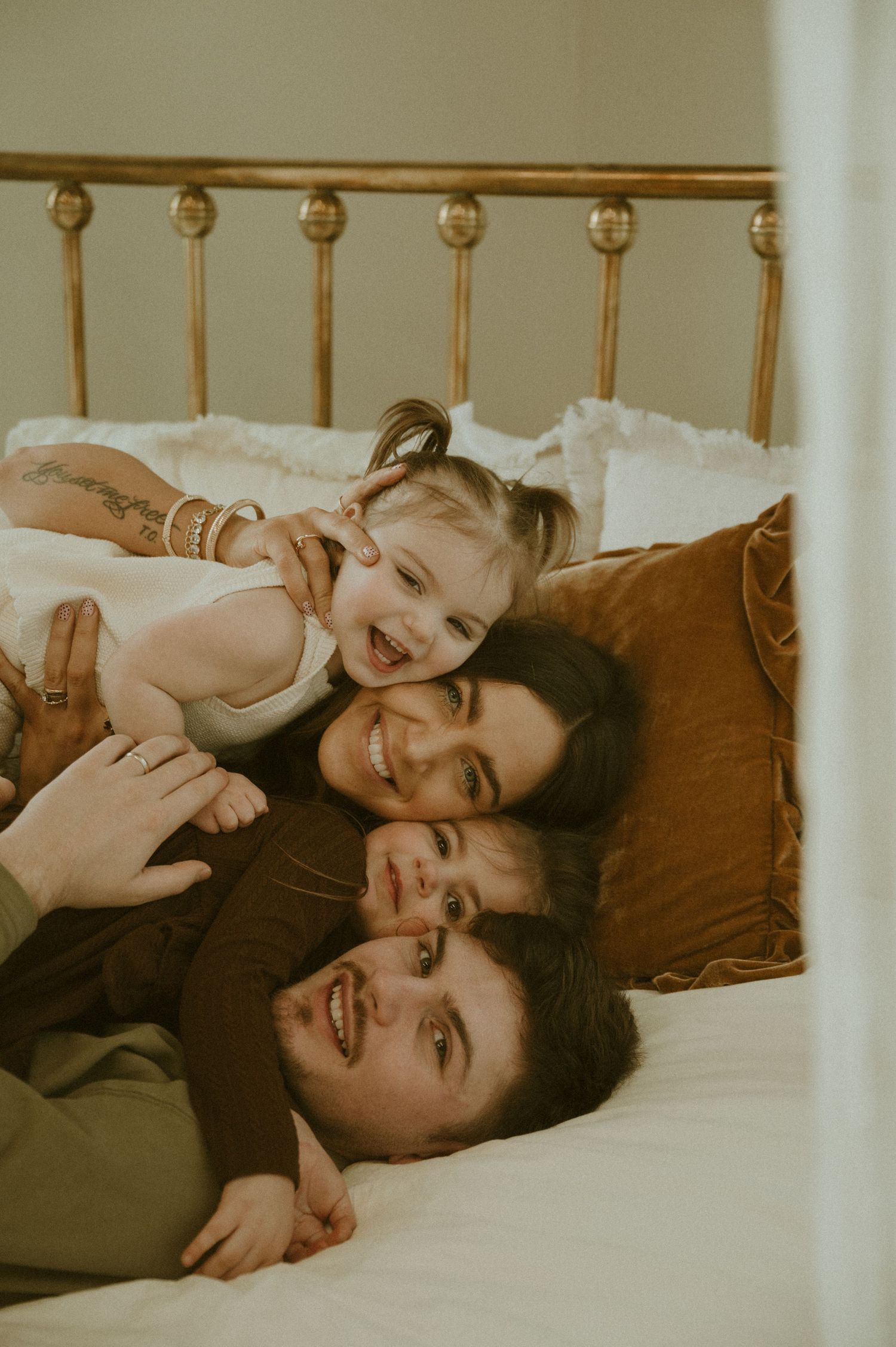 A family snuggles together on a bed with brown pillows in a warm-toned intimate portrait.