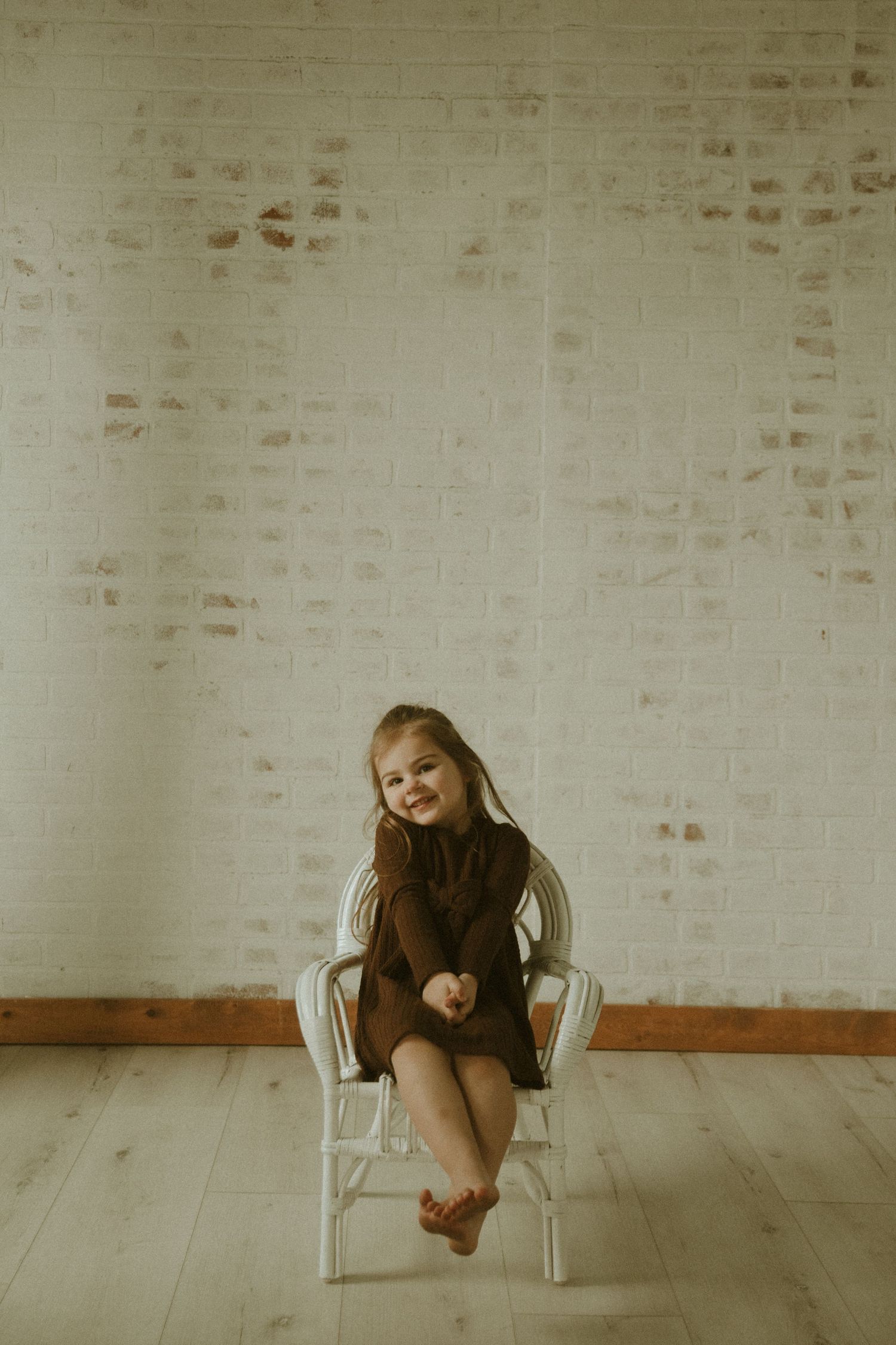 A person in brown clothing sits and poses playfully in a white chair against a light brick wall.