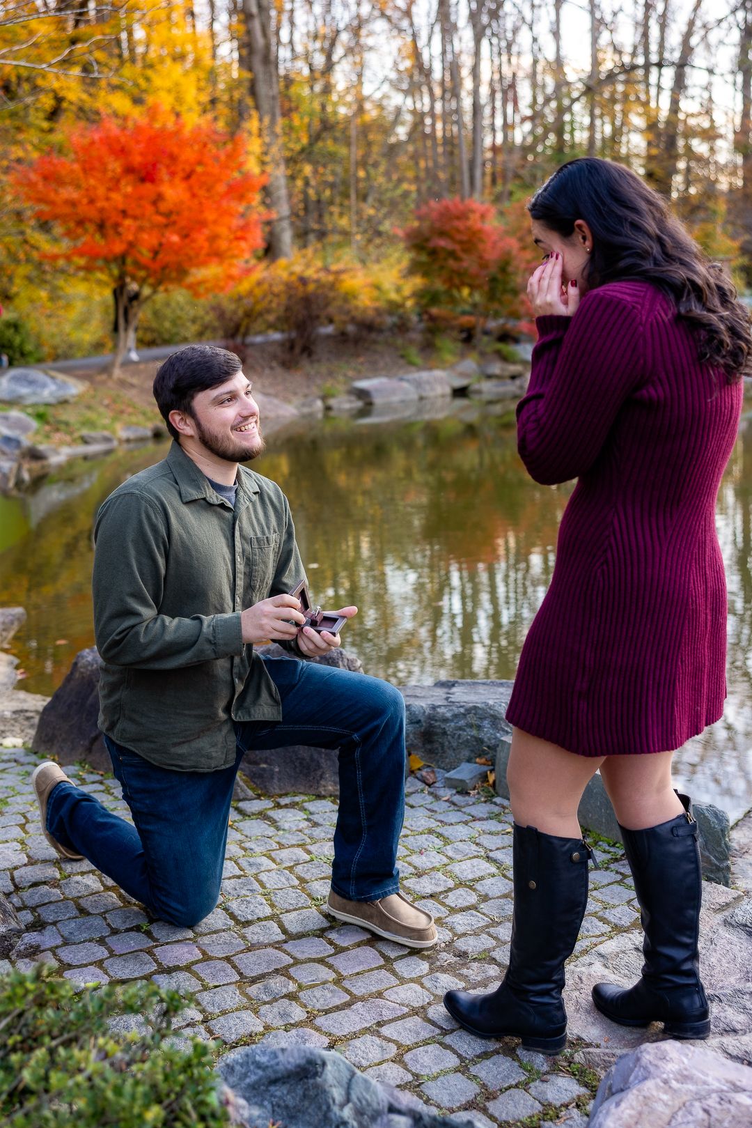 A romantic autumn proposal scene unfolds by a tranquil pond surrounded by vibrant fall foliage.
