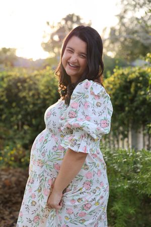 A person wearing a floral print dress poses in a sunny garden setting with greenery in the background.