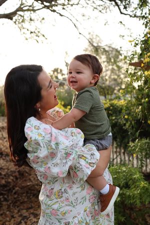 A woman in a floral dress lifts and plays with a small child wearing green in a sunlit garden setting.