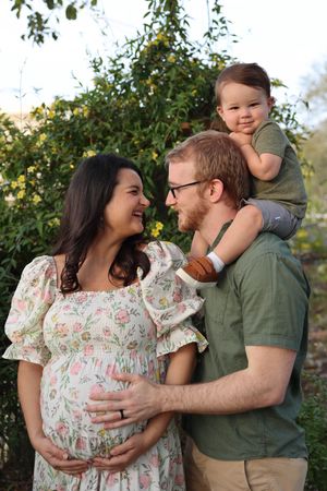 A couple shares tender moments in a garden setting wearing floral dress and green shirt while holding a small child.