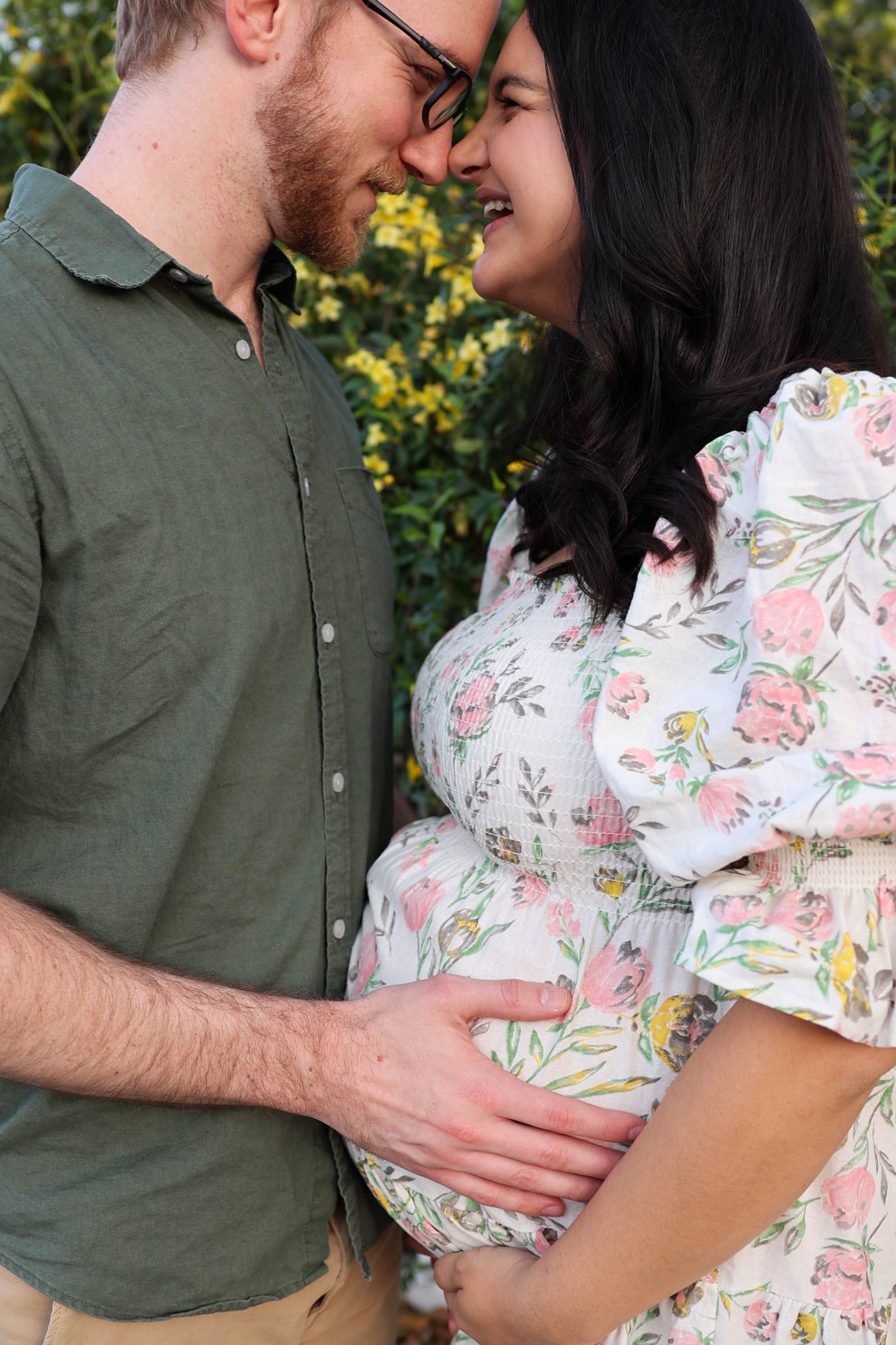 A couple in green and floral print outfits share a tender moment during a maternity photo shoot in a garden setting.