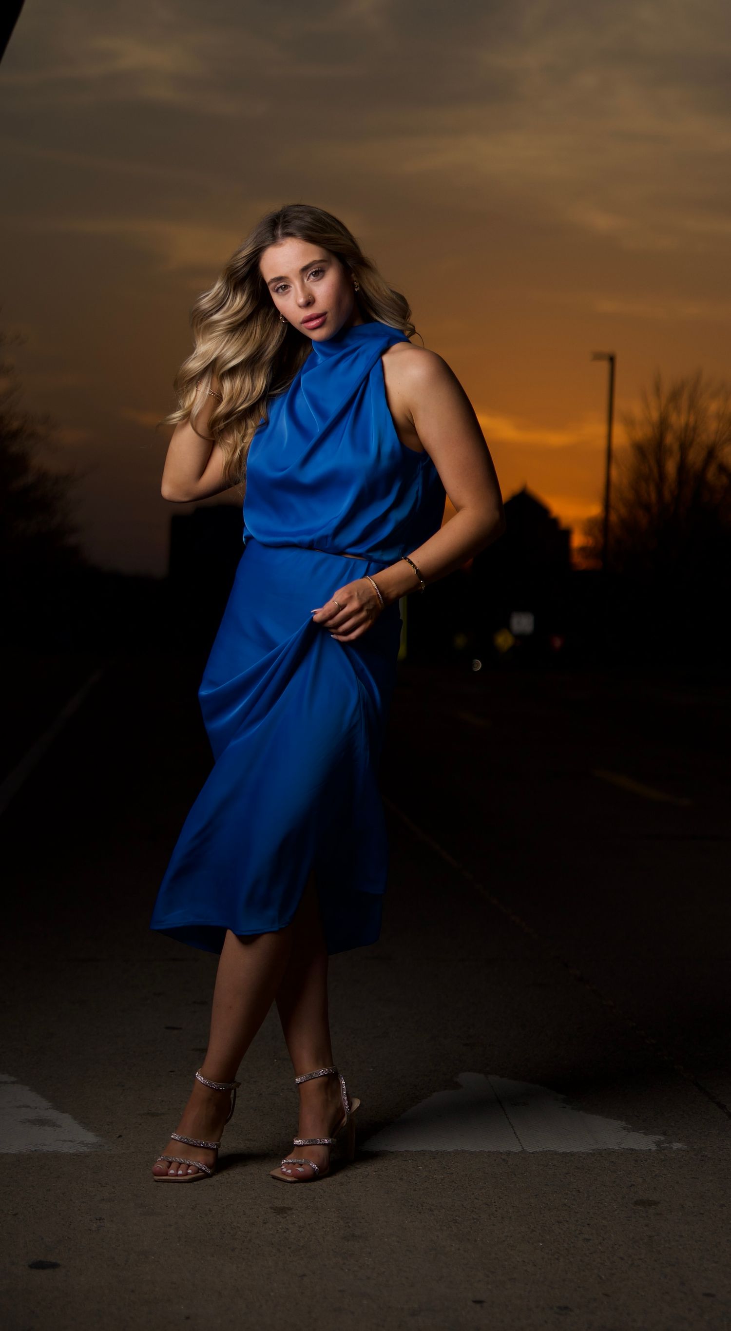 A person in a flowing blue dress poses dramatically against a sunset sky in a series of artistic dance-like movements.