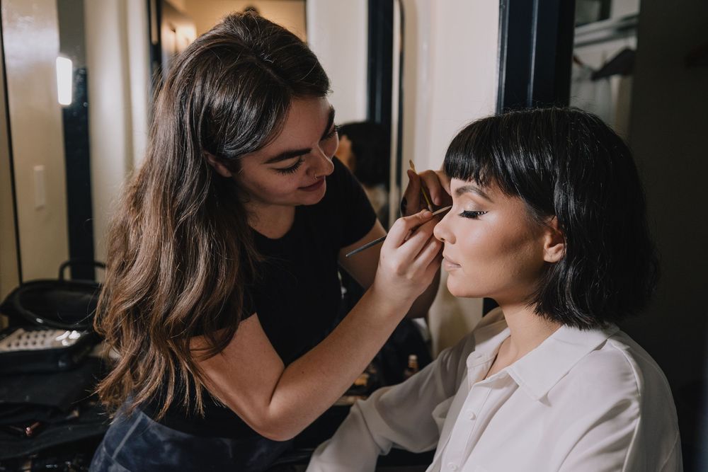 A professional makeup artist applying eyeshadow to a young woman in a dimly lit salon setting.