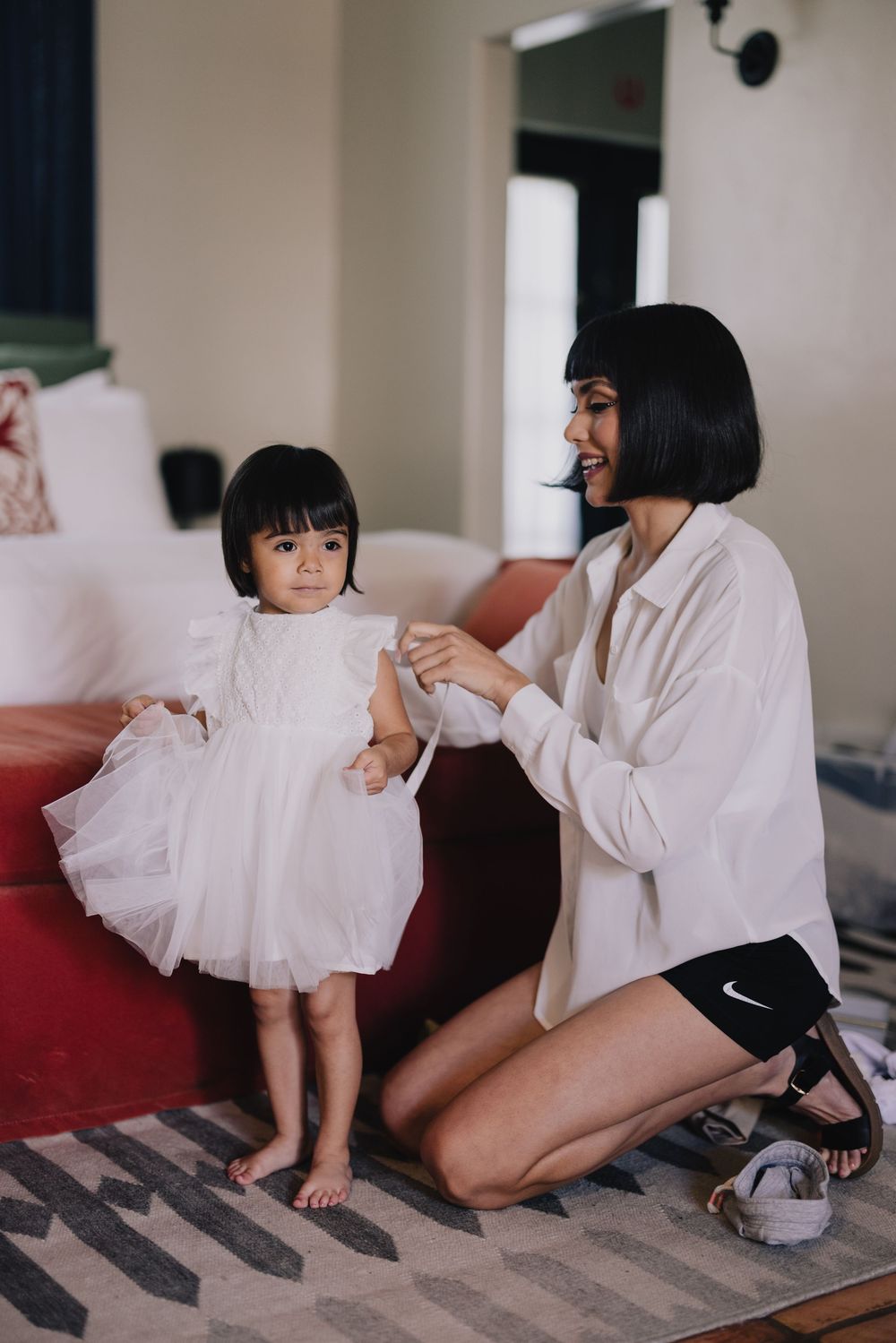 A mother kneels on a rug helping her toddler daughter in a white tutu dress stand in a cozy living room.