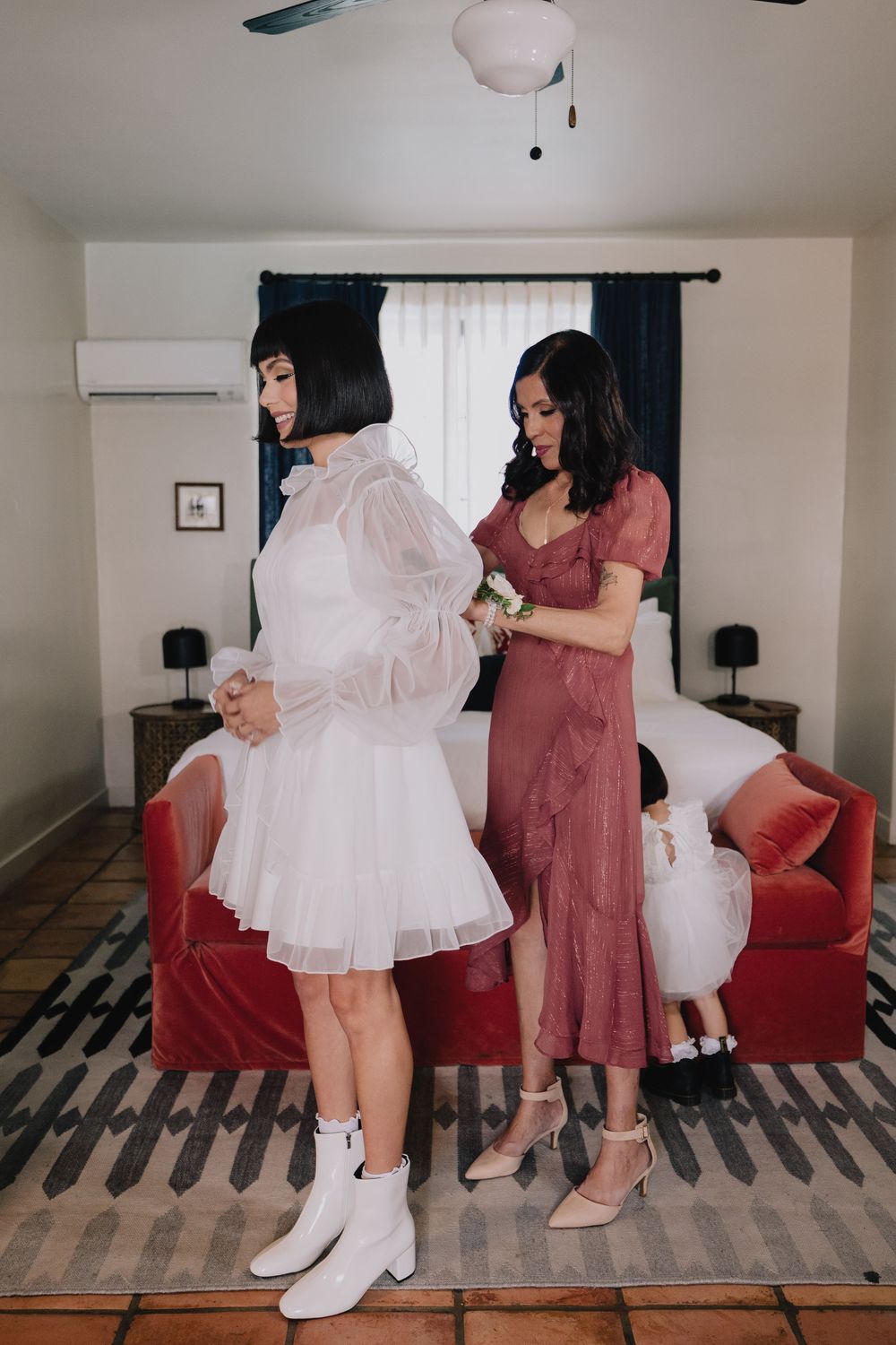 Bride in white mini dress with puff sleeves stands as bridesmaid in rose gown helps with bouquet in elegant room.