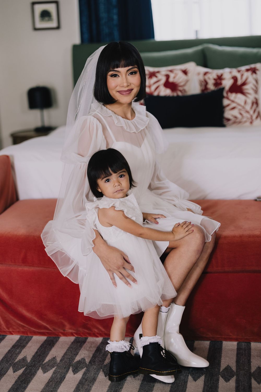 A bride in a short white tulle dress and veil sits on a red sofa holding a young girl in a matching white dress.