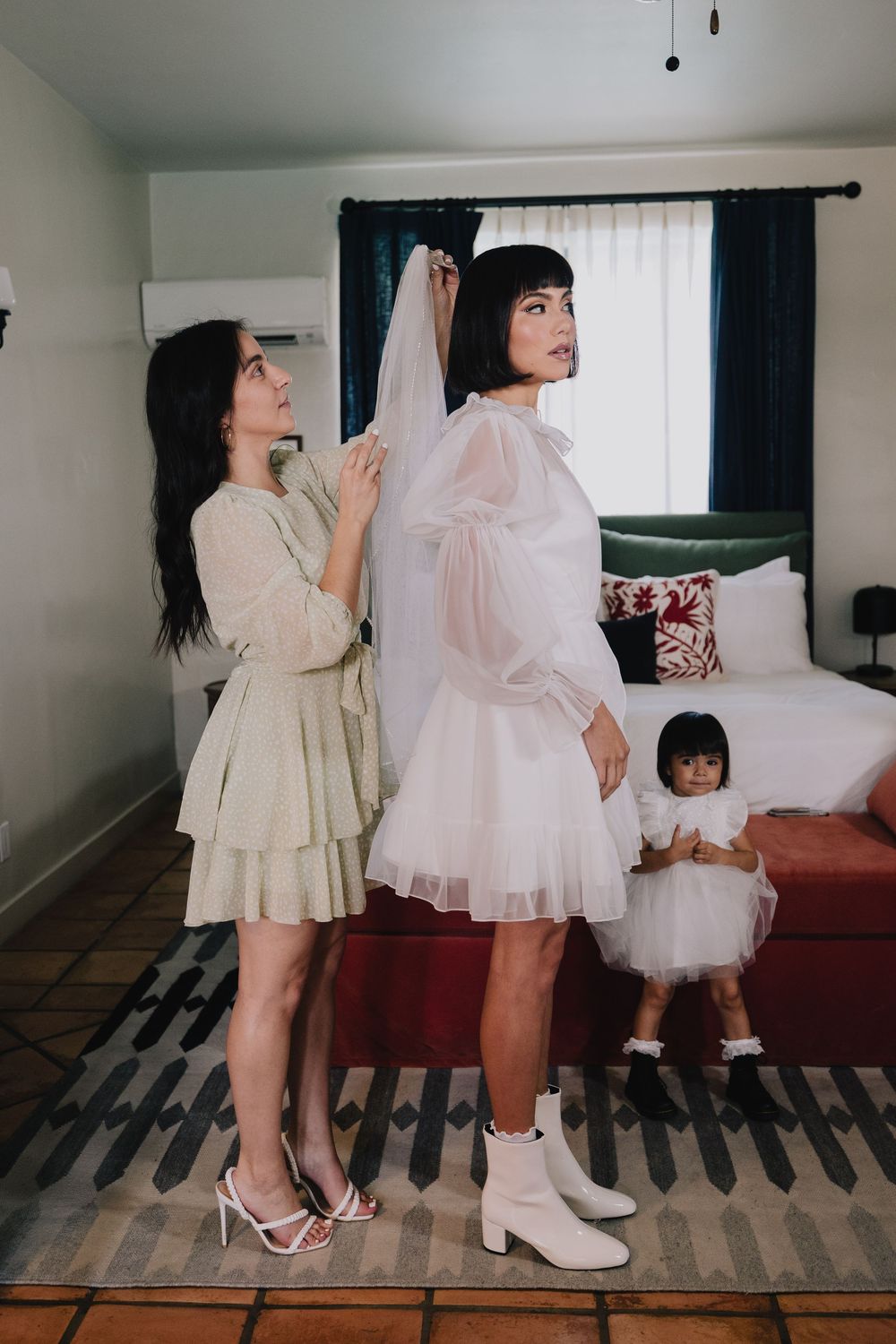 A bridesmaid in green adjusts the veil of a bride in a short white dress as a little girl watches in a hotel room.