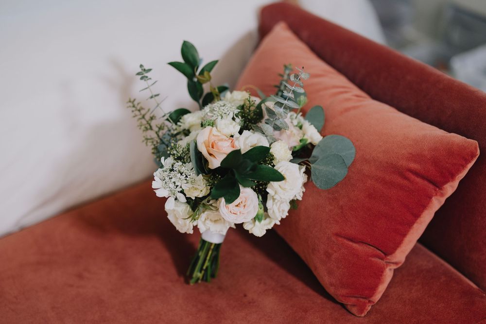 A bridal bouquet of white and blush roses with eucalyptus rests on a rust velvet pillow.