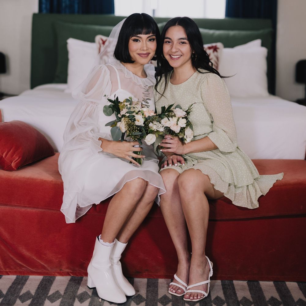 Two women in elegant dresses holding bouquets sit together on a red velvet sofa in a stylish hotel room.