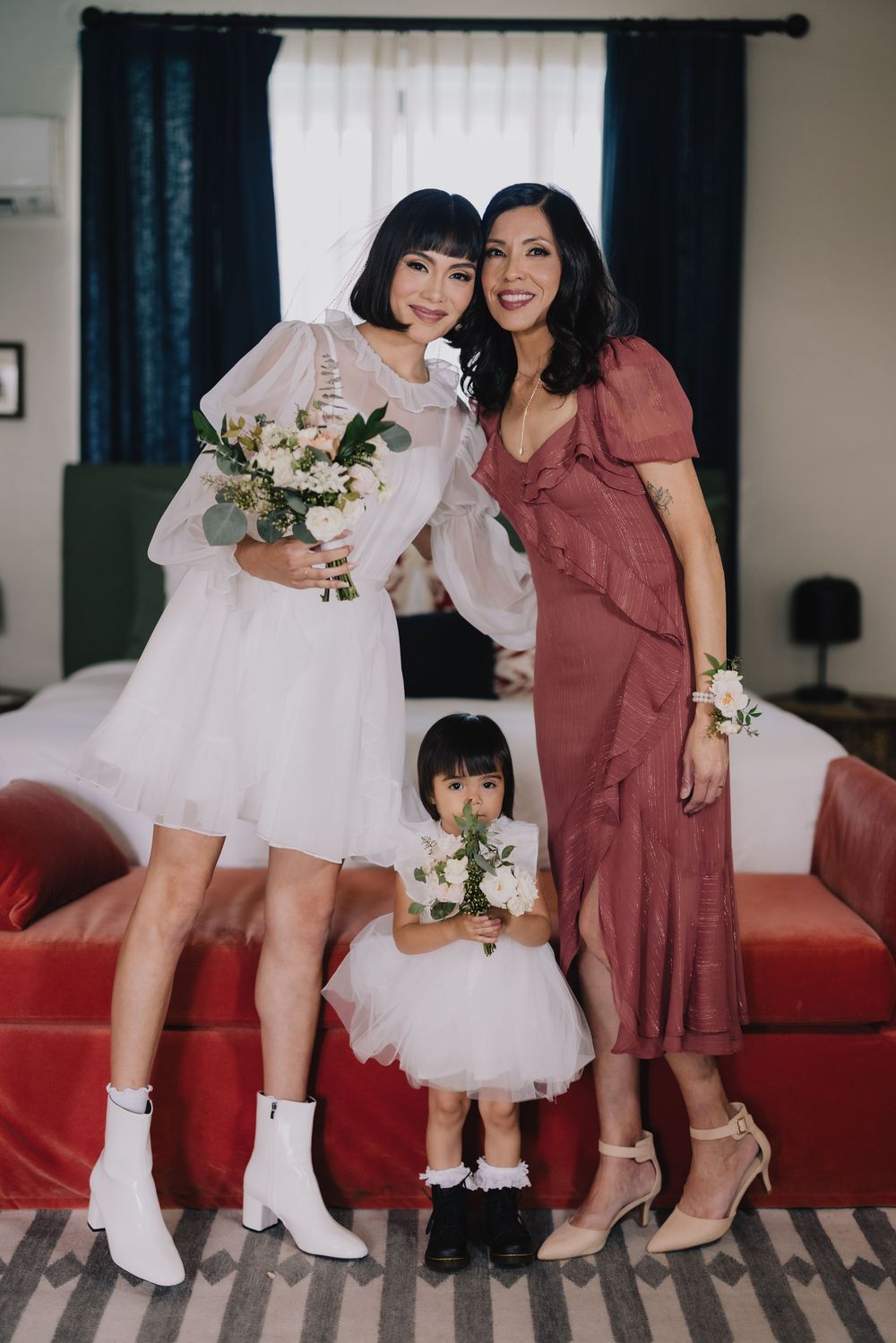 Bride in white dress and boots poses with bridesmaid in rose dress and flower girl holding bouquets indoors.