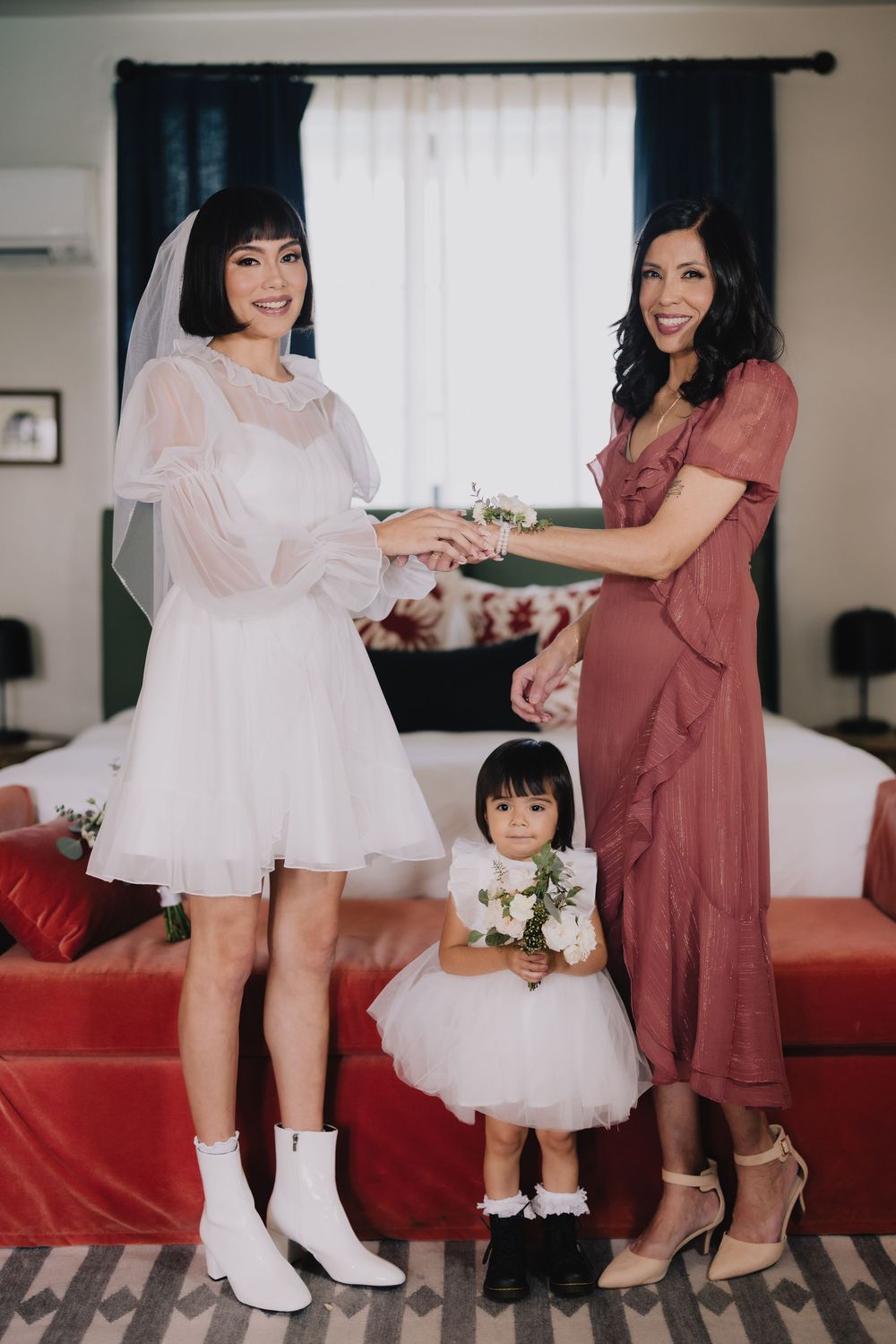 Bride in short white dress and veil with bridesmaid in dusty rose and flower girl holding white bouquet indoors.