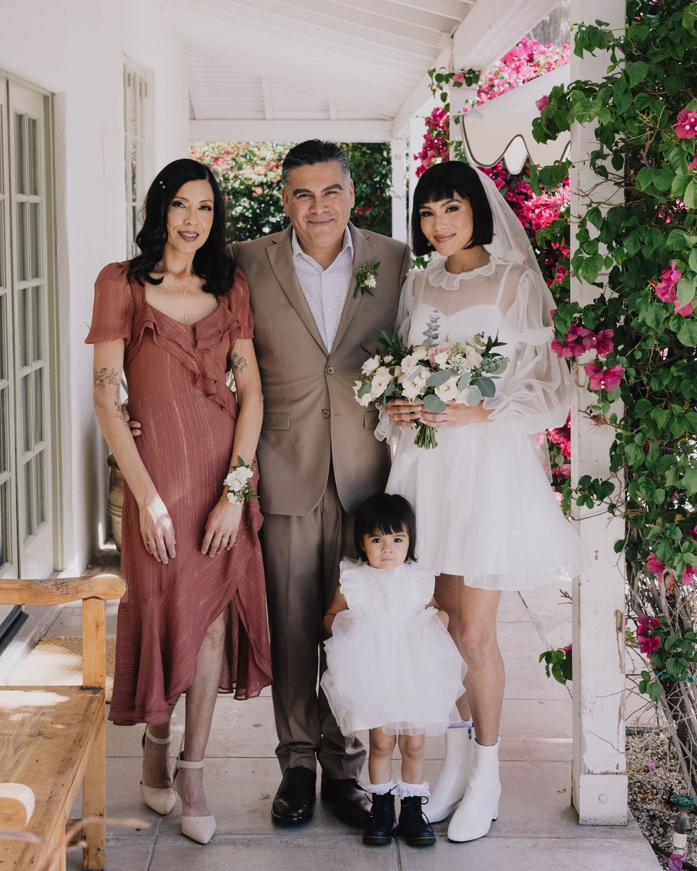 Family portrait at a wedding with bride in white mini dress, groom in tan suit, woman in pink dress, and flower girl.
