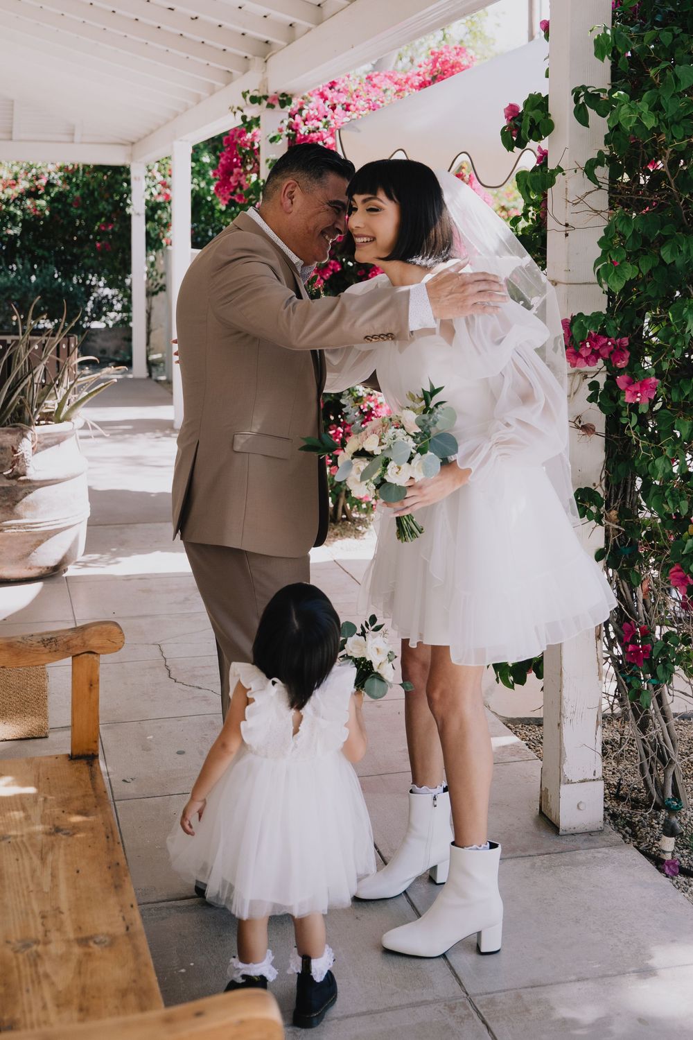 Groom in tan suit embracing bride in short white dress and boots at outdoor wedding ceremony with pink flowers.
