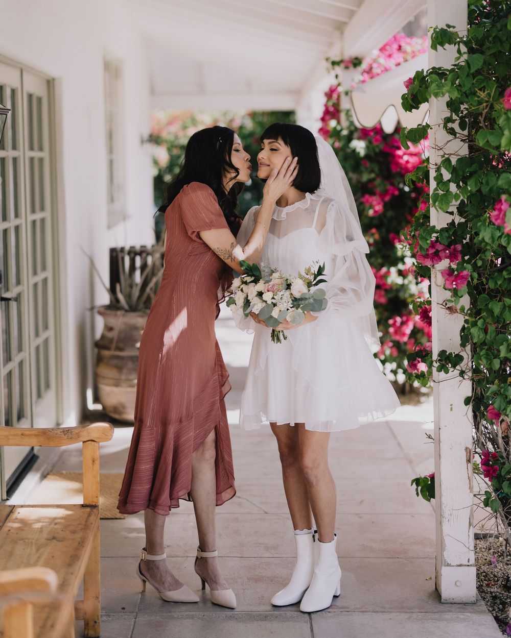 Two women share a tender moment outdoors, one in a white wedding dress with veil holding a bouquet.