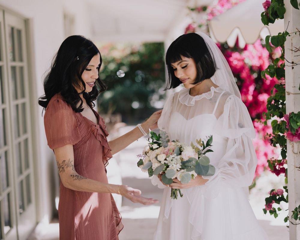 Bride in white gown holding bouquet with bridesmaid in pink dress at elegant outdoor wedding venue.