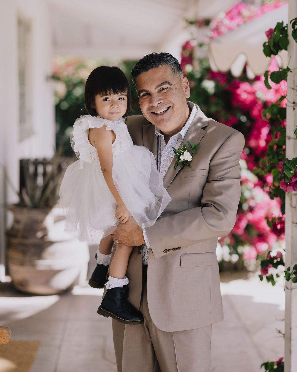 Man in tan suit holding young girl in white dress at outdoor wedding with pink bougainvillea flowers.