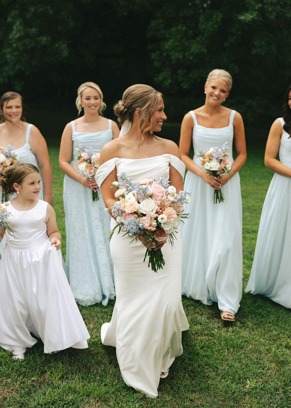 Bride in off-shoulder gown holds floral bouquet surrounded by bridesmaids in light blue dresses and flower girl outdoors.