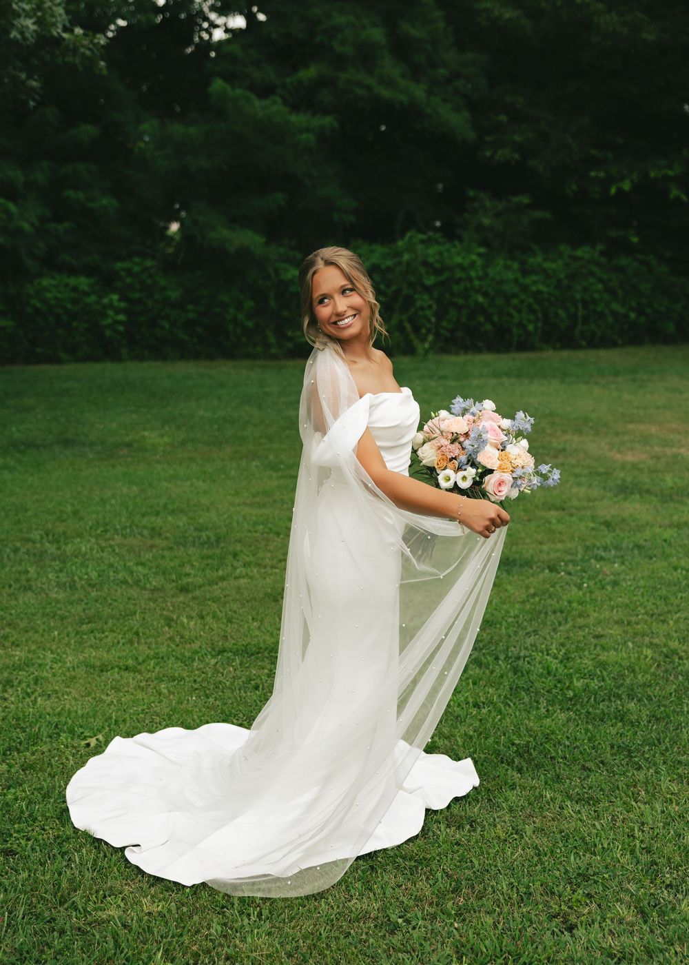 Smiling bride in off-shoulder white gown with flowing veil holds colorful bouquet on green lawn.