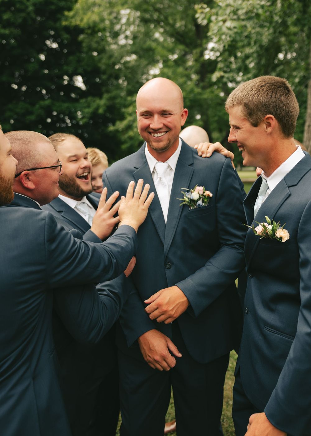 Groom in navy suit laughing with groomsmen outdoors, wearing floral boutonniere at wedding celebration.