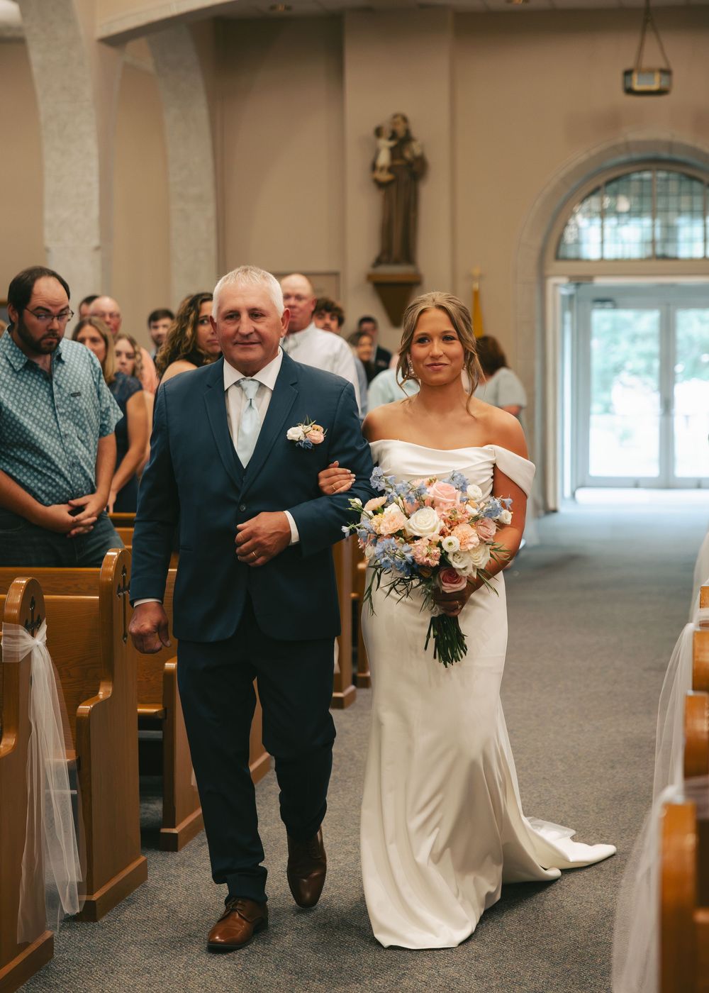 Bride in white gown walks down church aisle with escort holding pink and blue bouquet at wedding ceremony.