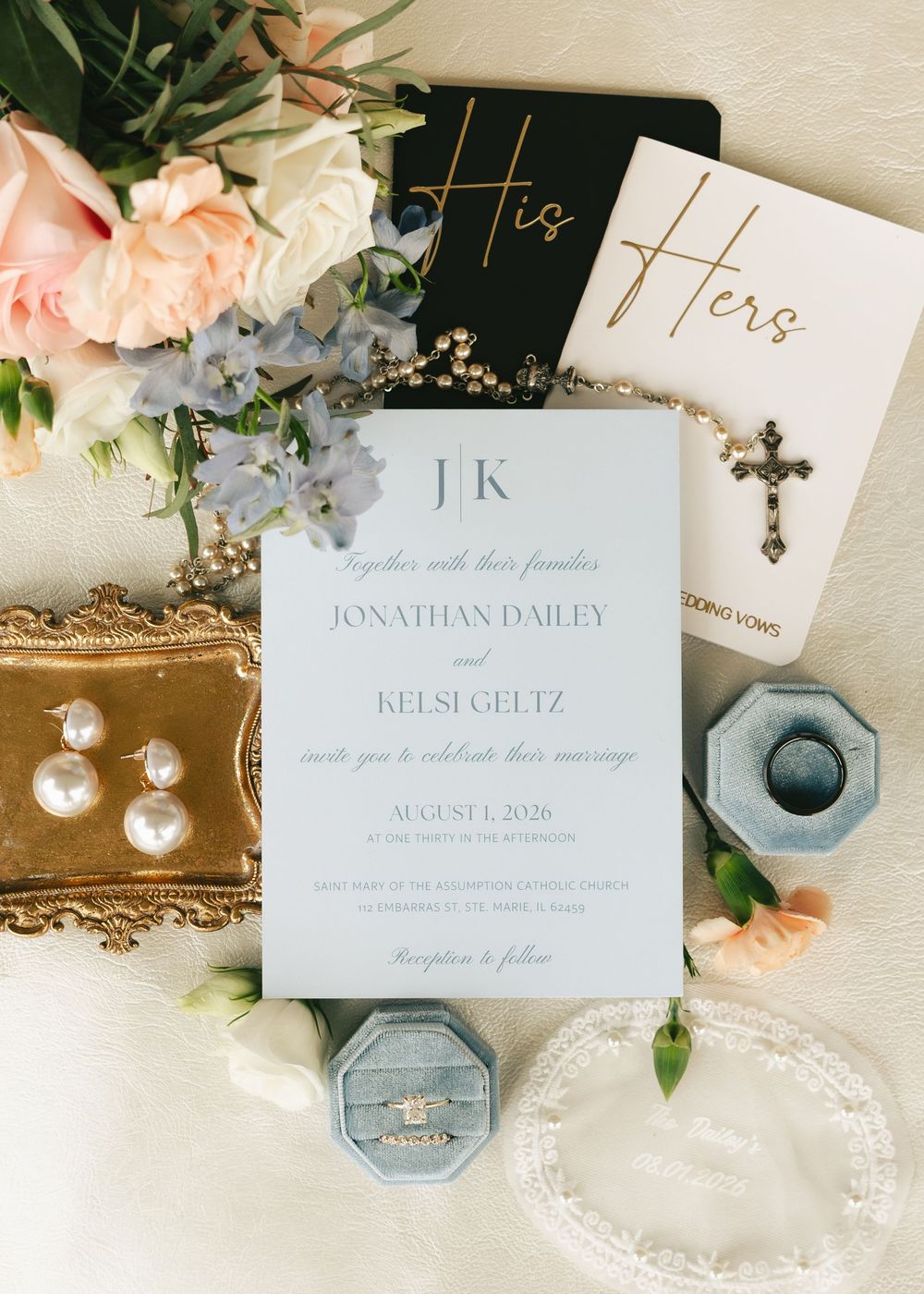 Elegant wedding flat lay with dusty blue invitation suite, pearl earrings, cross necklace, ring box, and blush floral bouquet.