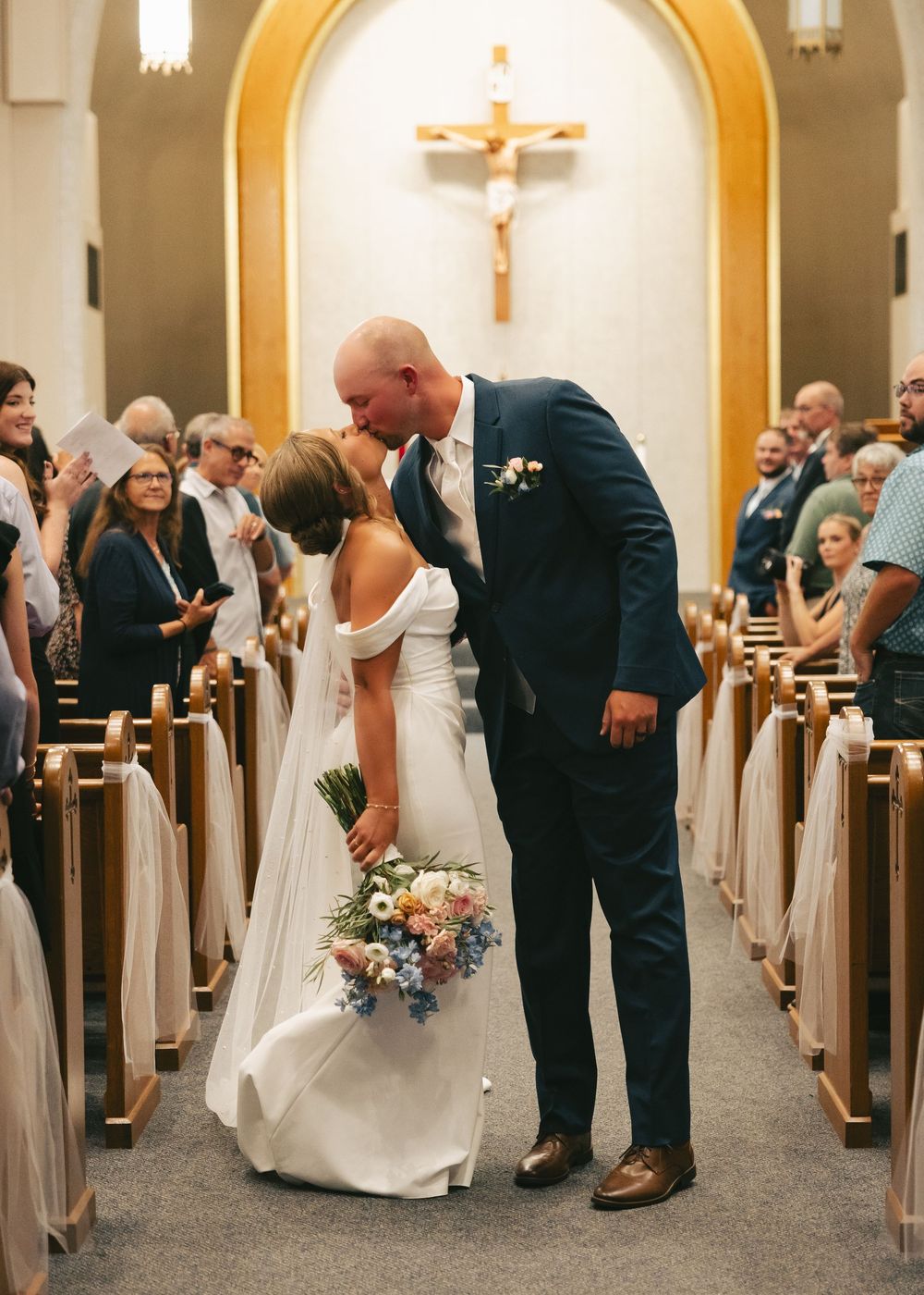 Bride and groom share a kiss in the church aisle after their wedding ceremony, with guests seated in wooden pews.