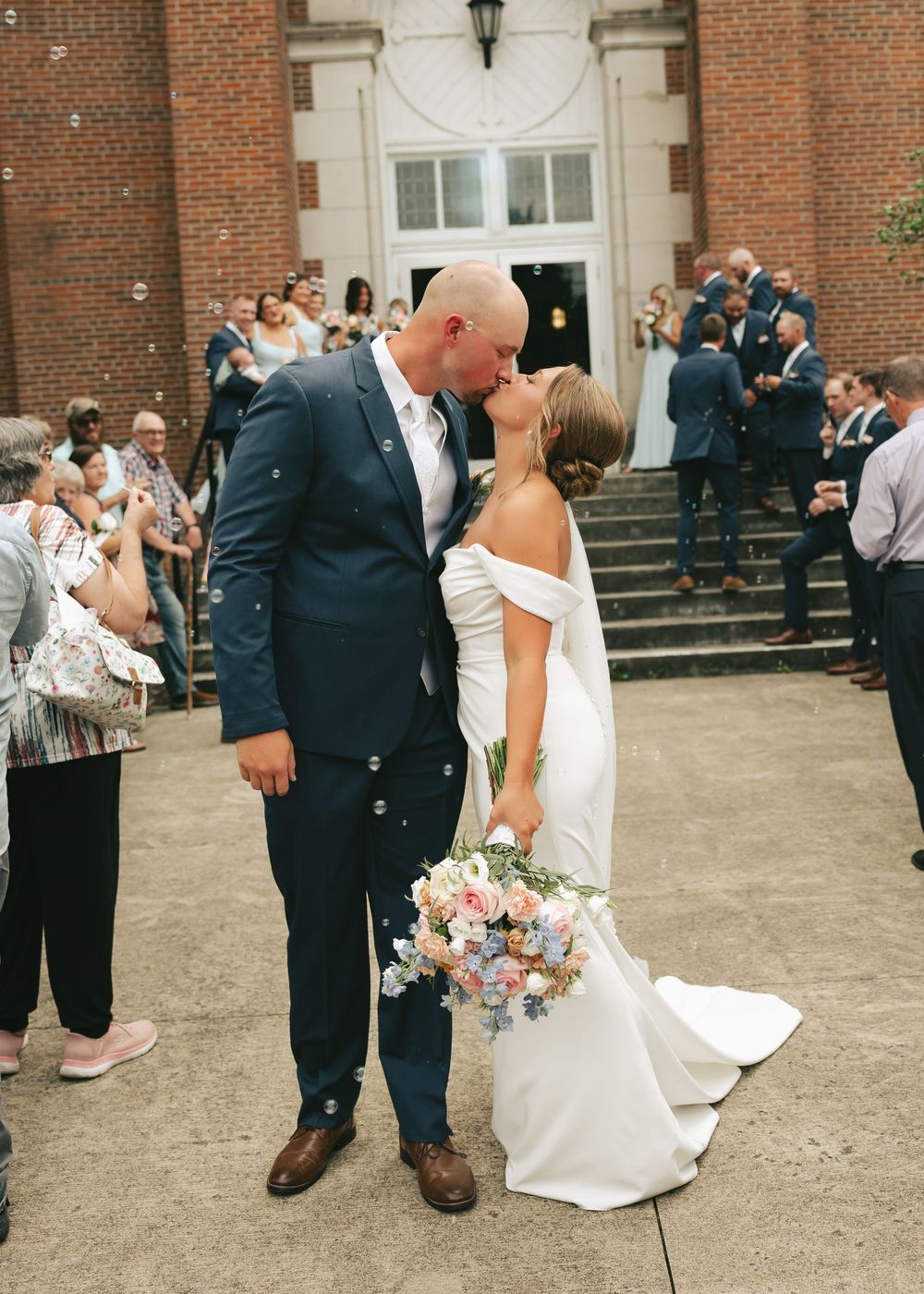 Newlyweds share a kiss outside brick venue, bride holding pastel bouquet, guests celebrating around them.